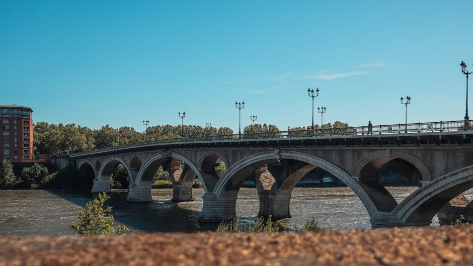 A stone bridge with multiple arches and decorative lamps along its railing spans a wide river in the daytime. The river has a gentle flow, with visible ripples and some small trees or bushes growing along its edge. In the background, there are trees, a red brick building, and a clear blue sky with a few wispy clouds. The scene is outdoors, on a bright day, with the bridge likely used for pedestrian and vehicle traffic. Although no specific interior or moving process is visible, this setting suggests a location related to house removals or furniture transport between nearby areas, possibly part of a home relocation service offered by Man with Van Castelnau.