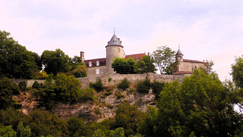 A historic stone castle with multiple conical-roofed turrets and narrow windows, situated at the end of a straight gravel pathway leading through a well-maintained grassy lawn. The castle’s exterior