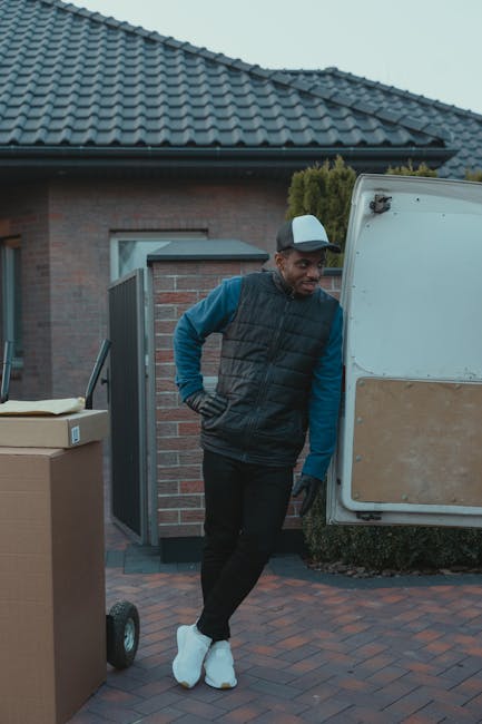 A man dressed in a black vest, blue long-sleeve shirt, black pants, and white footwear leaning against the side of a white moving van with a wooden panel on its exterior, parked outside a residential 