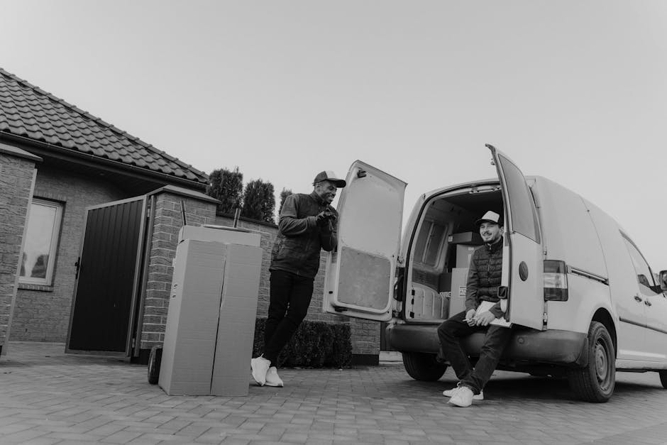 A man dressed in a black vest, blue long-sleeve shirt, black pants, and white footwear leaning against the side of a white moving van with a wooden panel on its exterior, parked outside a residential 