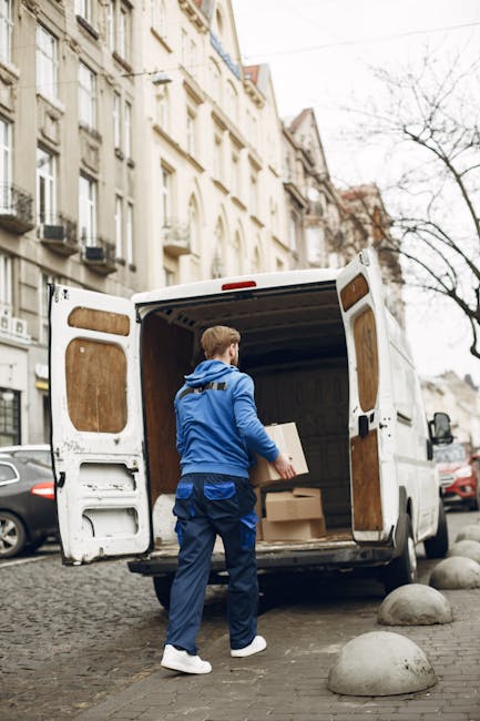 A professional mover wearing a dark vest and cap is loading cardboard boxes into the open rear doors of a white cargo van parked outside a modern building with large glass windows. The boxes are vario