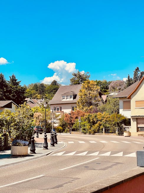A quiet residential street in Castelnau during daytime, showing a row of houses with pitched roofs and a mix of brick and stucco exteriors, surrounded by trees with green and autumnal foliage. The sky is bright blue with scattered white clouds, and the street is lined with black bollards and planters filled with flowers. In the foreground, a man with a van from Man with Van Castelnau is engaged in the loading process, carrying large cardboard boxes and wrapped furniture pieces towards the vehicle parked on the pavement. The van is partially visible, positioned near the curb, with its rear doors open. The scene captures the activity involved in home relocation, including furniture transport and careful packing, with evidence of the logistics involved in a professional removals service for house moves in the SW13 area.