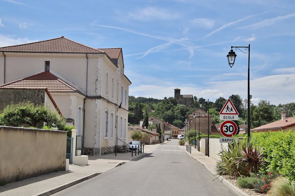 A quiet residential street in Castelnau featuring a cream-colored, two-storey house with a tiled roof on the left side, partially bordered by a low wall and greenery. The pavement runs along the front of the house, with a white vehicle parked near the curb. On the right side, there is a black street lamp with a decorative arm, adjacent to a red and white triangular road sign indicating school children and a circular sign displaying a speed limit of 30 mph, with the words 'École' and 'Rappel' written on them. The street extends into the distance with more houses, trees, and a hilltop building visible against a partly cloudy blue sky, suggesting a suburban environment. This scene captures an early stage of home relocation or furniture transport process, with the quiet street ready for a moving company's arrival, possibly associated with Man with Van Castelnau, as part of packing and loading activities for house removals.