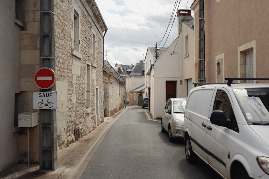A quiet residential street in Castelnau during daytime, showing a row of houses with pitched roofs and a mix of brick and stucco exteriors, surrounded by trees with green and autumnal foliage. The sky is bright blue with scattered white clouds, and the street is lined with black bollards and planters filled with flowers. In the foreground, a man with a van from Man with Van Castelnau is engaged in the loading process, carrying large cardboard boxes and wrapped furniture pieces towards the vehicle parked on the pavement. The van is partially visible, positioned near the curb, with its rear doors open. The scene captures the activity involved in home relocation, including furniture transport and careful packing, with evidence of the logistics involved in a professional removals service for house moves in the SW13 area.