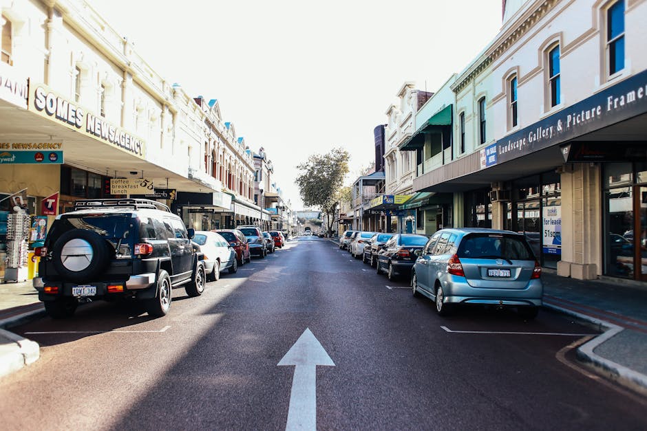 A straight, two-way urban street in Castelnau with parked cars lining both sides, including a black SUV on the left and a silver hatchback on the right. The street runs between commercial buildings with various storefronts, shop signs, and awnings visible above their entrances. There is a clear white arrow painted on the tarmac indicating the direction of traffic flow. The road is surfaced with smooth asphalt, and the weather appears sunny with bright lighting and minimal shadows cast by the buildings and vehicles. No active loading or moving process is visible in this image, but the scene depicts a typical location where home relocation or furniture transport activities could occur, especially considering the proximity of shops and parking spaces suitable for brief loading and unloading activities nearby. The image is focused on the street, which could be part of a related moving or packing and moving service discussed on the webpage about Packing and Parking on Lonsdale Road, Castelnau (SW13). Man with Van Castelnau is the local company serving such relocation needs.