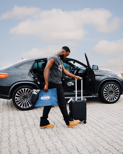 A man with dark skin, short curly hair, wearing a sleeveless grey hoodie, black pants, and tan work boots, is standing next to a black luxury sedan on a paved parking area. He is holding a black rolling suitcase in one hand and a blue cardboard box with a white logo in the other hand. The car's front passenger door, which is open, reveals the interior, while the trunk area is partly visible in the background. On the ground beside him is a small black carry-on bag. The scene is outdoors under a partly cloudy sky, likely during daylight hours, and the environment suggests a home relocation or moving process serviced by [COMPANY_NAME], a professional removals company specializing in packing and moving logistics, including furniture transport and vehicle loading. The image captures the process of loading belongings into the vehicle for a home move or packing and moving task.