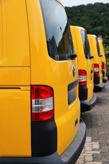 A row of three yellow vans parked on a paved street during daylight hours, with the rear ends visible and the windows reflecting nearby trees and buildings. The vans are positioned closely together, facing away from the camera, with their black rear bumpers and red and white rear lights clearly shown. The scene is set on Castelnau Road, with a background of lush green trees on a hillside, indicating a residential or suburban area. The image captures the exterior of the vans used in home relocation or furniture transport services, reflecting the logistics involved in a professional removal process, as provided by Man with Van Castelnau, during a typical loading or parking scenario in the context of house removals and moving logistics.