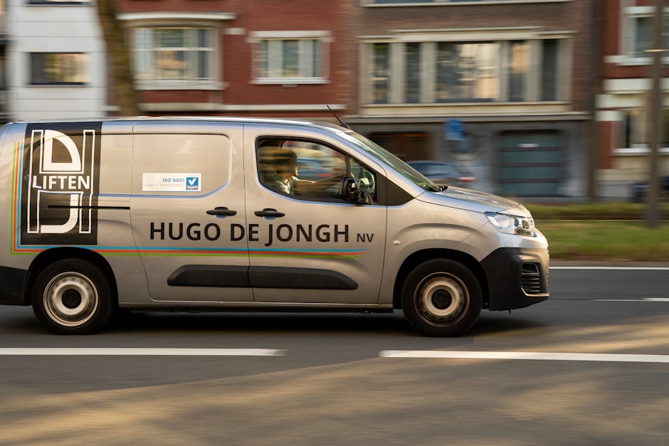 A grey commercial van belonging to Hugo De Jongh NV is parked on the street in front of residential buildings with multiple stories. The van is labeled with the company logo and name, and features a black front bumper, side mirrors, and black wheel arches. The vehicle's windows show reflections of the surroundings and a driver inside. The van appears to be in motion, with a blurred background of multi-storey brick and concrete apartment buildings, some with balconies, and a few cars traveling in the opposite direction. On the pavement, a small grassy area separates the sidewalk from the road, with a lamppost visible on the right side of the image. The scene is lit by natural daylight, providing clear visibility of the vehicle and urban environment, which aligns with house removals and furniture transport logistics managed by Man with Van Castelnau during a typical home relocation process.