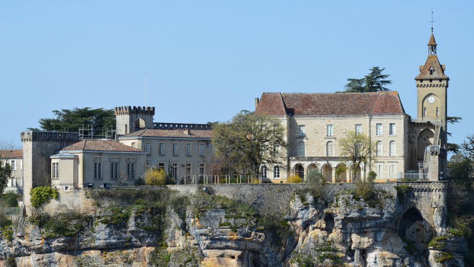 No objects related to house removals or moving services are visible in this image. The photograph depicts a historic stone building with a tower, located on a rocky hillside, surrounded by trees and foliage under a clear blue sky. The building features a mix of architectural elements, including arched windows and a clock tower. There are no furniture, boxes, packaging materials, or transportation vehicles present. The scene appears to be taken from a distance, showing the entire structure and its natural environment, without any visible activity such as loading, carrying, or packing, consistent with a static, exterior view.