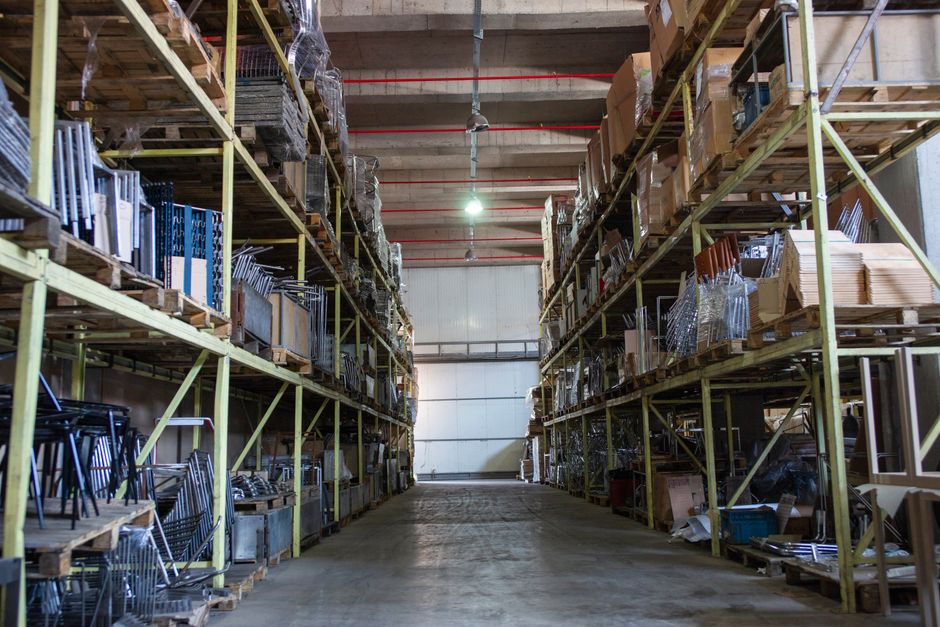 Inside a spacious warehouse with high ceilings and industrial lighting, two sets of tall yellow metal shelving units extend from the foreground to the back wall, stocked with various household items and furniture parts. On the left shelving, there are stacks of cardboard boxes, some wrapped in plastic, along with metal chairs and small furniture pieces arranged on different levels. The right shelving contains wooden panels, plastic-wrapped items, and additional boxes, all organized systematically. The concrete floor is clear, providing space for movement during the loading or packing process. A large, closed white sectional garage door is visible at the end of the aisle, with a bright ceiling light illuminating the scene. The environment suggests a storage or staging area used during home relocation and furniture transport, with visible packaging materials like cardboard and plastic to protect items during packing and moving. Man with Van Castelnau uses such facilities to assist with their removals and short-term storage solutions.