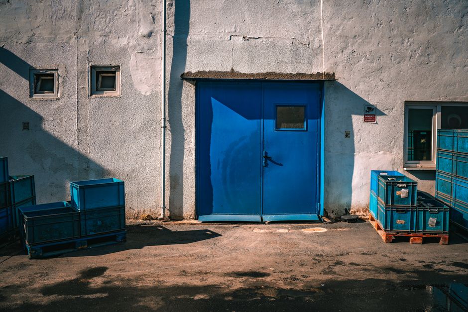 The image shows the exterior of a building with a large blue metal door situated in the centre, secured with a handle and lock, and a small rectangular window near the top. The building’s white textured wall has three small square windows, two on the left and one on the right of the door. To the right of the blue door, there is a small metal sign, and a larger window behind a metal railing. On the ground, adjacent to the door, there are four blue plastic containers stacked on wooden pallets, used for packing or storage, positioned on a paved surface with shadows cast by nearby sunlight. The scene appears to be outside a commercial or industrial property, capturing a moment in a home relocation or moving process with a focus on prepared materials and basic loading arrangements. Man with Van Castelnau, a professional removals service, could be involved in managing the transportation or storage of household belongings during relocations, as suggested by the context of storage solutions and logistical considerations.
