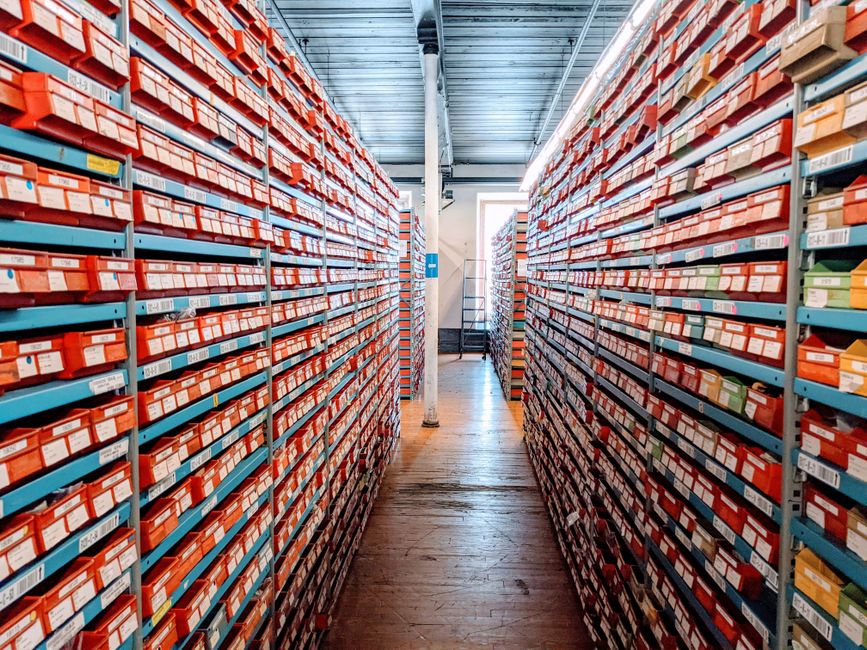 Inside a spacious warehouse with high ceilings and industrial lighting, two sets of tall yellow metal shelving units extend from the foreground to the back wall, stocked with various household items and furniture parts. On the left shelving, there are stacks of cardboard boxes, some wrapped in plastic, along with metal chairs and small furniture pieces arranged on different levels. The right shelving contains wooden panels, plastic-wrapped items, and additional boxes, all organized systematically. The concrete floor is clear, providing space for movement during the loading or packing process. A large, closed white sectional garage door is visible at the end of the aisle, with a bright ceiling light illuminating the scene. The environment suggests a storage or staging area used during home relocation and furniture transport, with visible packaging materials like cardboard and plastic to protect items during packing and moving. Man with Van Castelnau uses such facilities to assist with their removals and short-term storage solutions.
